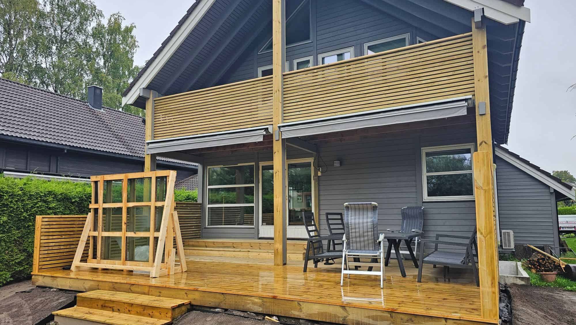Ny terrasse i tre med overbygg og møbler, sett mot moderne bolig med store vindusflater.